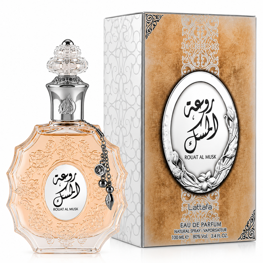 A bottle of Rouat Al Musk Eau de Parfum next to its retail box. The perfume bottle is clear glass filled with a pale peach-pink liquid, featuring an ornate silver-tone cap and a hanging silver charm.
